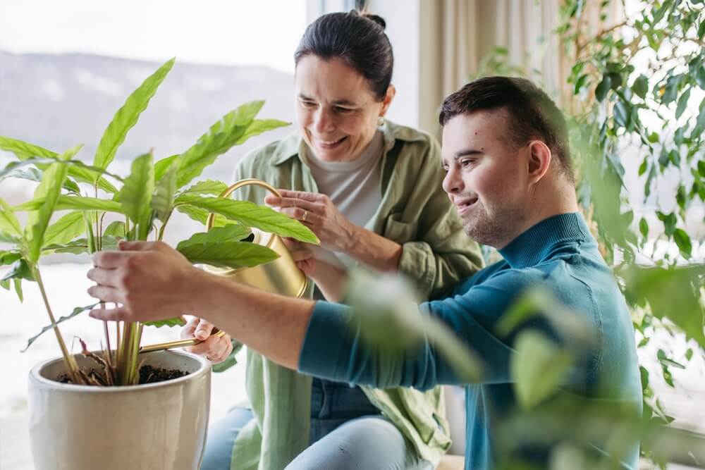 Young man with Down syndrome and his mother at home watering the plants.