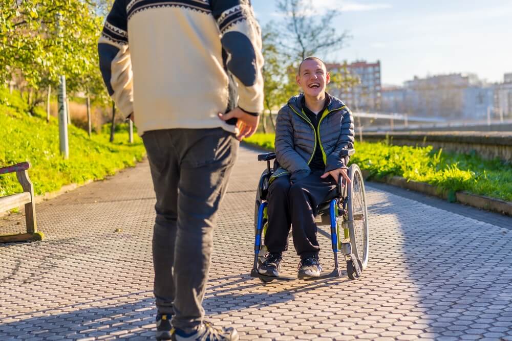 Disabled person in a wheelchair smiling with a family member taking a walk in a park.